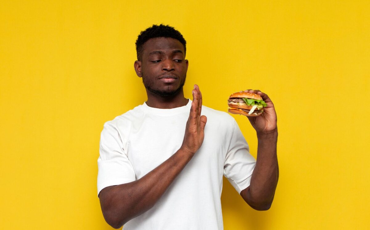 Photo of a man in front of a yellow background saying no to a burger, to illustrate article about a new study on what influences people to eat less meat