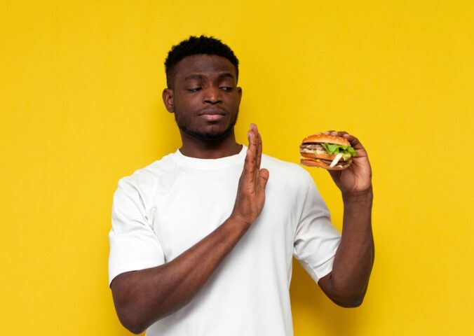 Photo of a man in front of a yellow background saying no to a burger, to illustrate article about a new study on what influences people to eat less meat