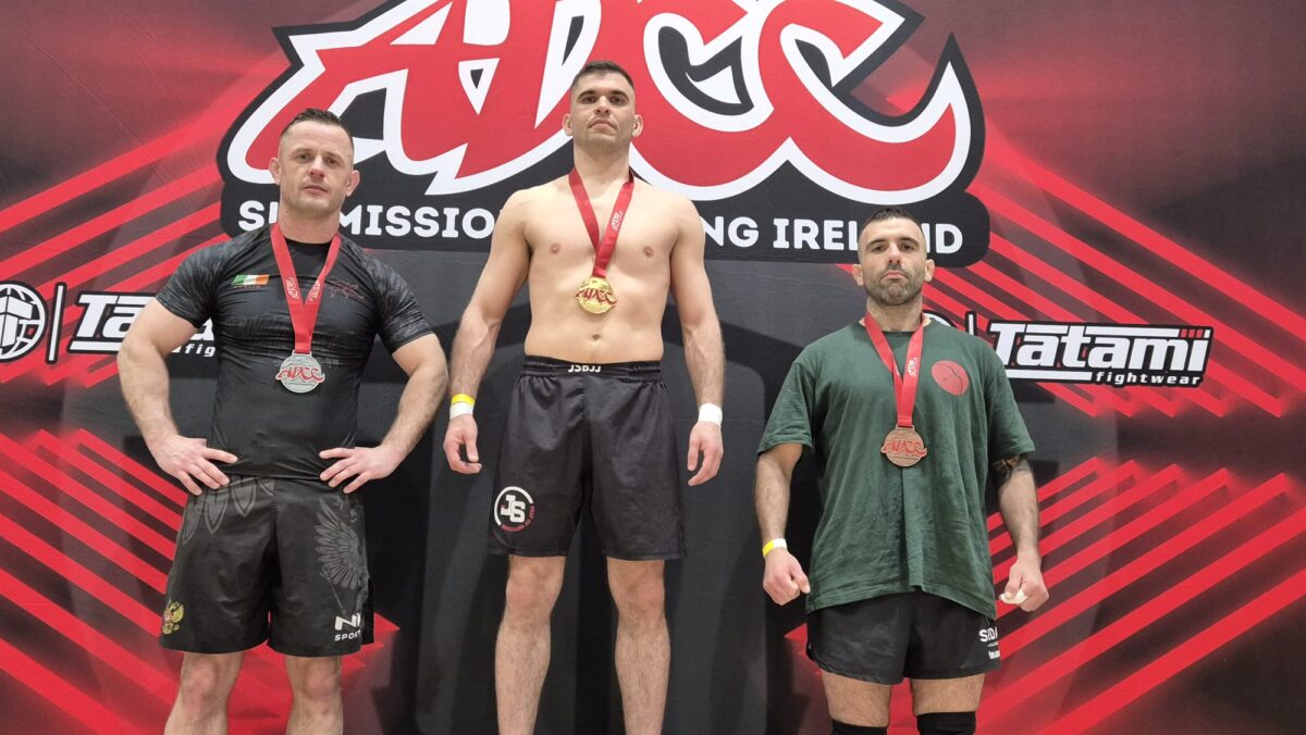 Photo shows Bernardo Ficarella, a vegan athlete and champion martial artist with black belts in both judo and Brazilian jiu-jitsu, on a podium and wearing a gold medal alongside two other athletes