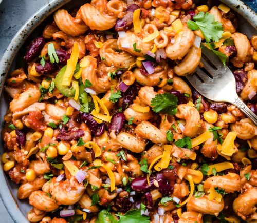 A vegan chili mac in a bowl next to dairy-free cheese and cilantro for basic vegan meals