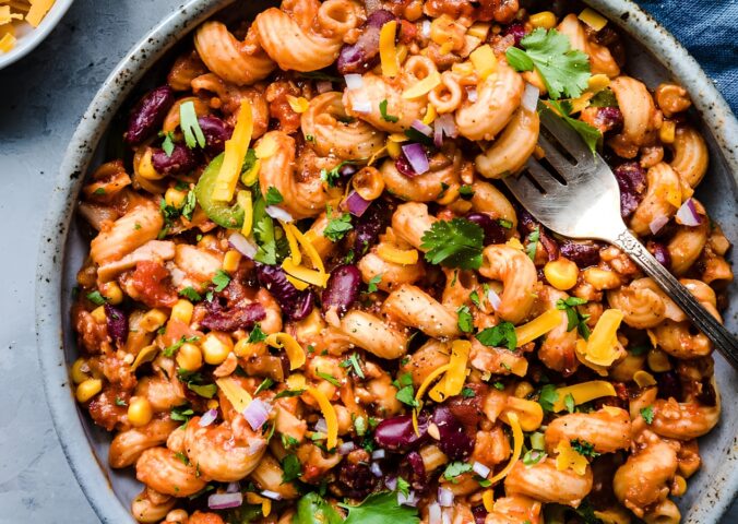 A vegan chili mac in a bowl next to dairy-free cheese and cilantro for basic vegan meals