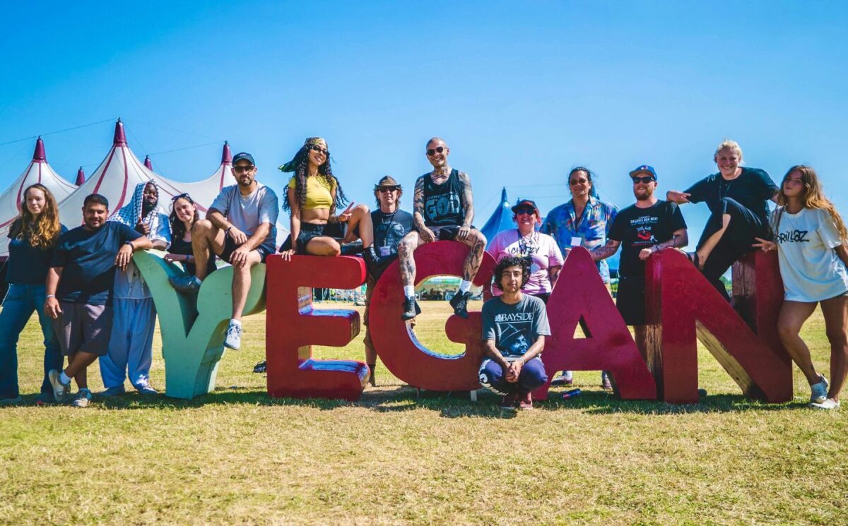 Photo shows the crew and organisers of Vegan Camp Out posing on a large "vegan" sign. Vegan Camp Out has announced the food lineup for this year’s festival