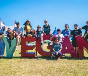 Photo shows the crew and organisers of Vegan Camp Out posing on a large "vegan" sign. Vegan Camp Out has announced the food lineup for this year’s festival