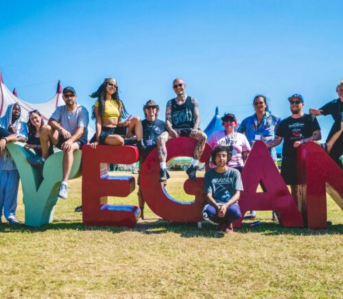 Photo shows the crew and organisers of Vegan Camp Out posing on a large "vegan" sign. Vegan Camp Out has announced the food lineup for this year’s festival