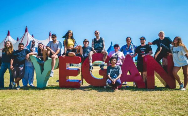 Photo shows the crew and organisers of Vegan Camp Out posing on a large "vegan" sign. Vegan Camp Out has announced the food lineup for this year’s festival