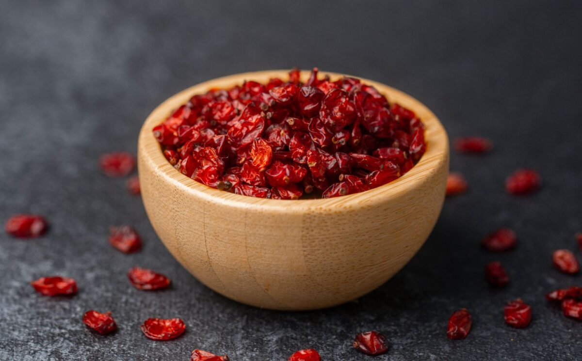 Dried barberries in a wooden bowl, with several berries spilled onto a black surface, to illustrate article about the berry that lowers cholesterol