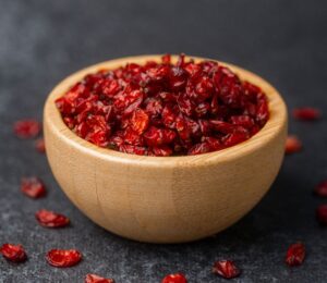 Dried barberries in a wooden bowl, with several berries spilled onto a black surface, to illustrate article about the berry that lowers cholesterol