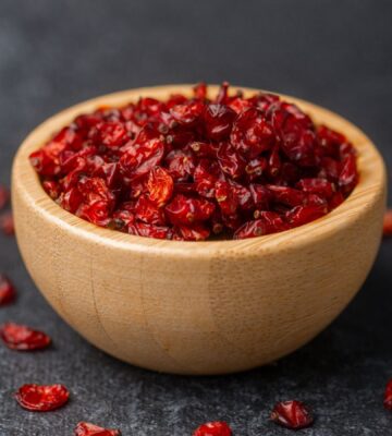 Dried barberries in a wooden bowl, with several berries spilled onto a black surface, to illustrate article about the berry that lowers cholesterol