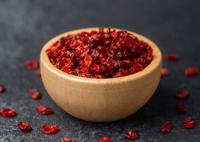 Dried barberries in a wooden bowl, with several berries spilled onto a black surface, to illustrate article about the berry that lowers cholesterol