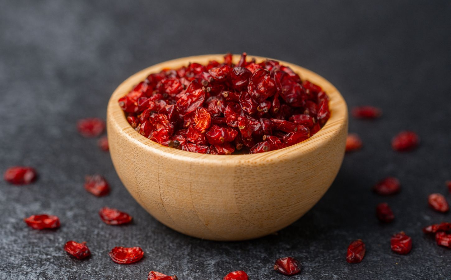 Dried barberries in a wooden bowl, with several berries spilled onto a black surface, to illustrate article about the berry that lowers cholesterol