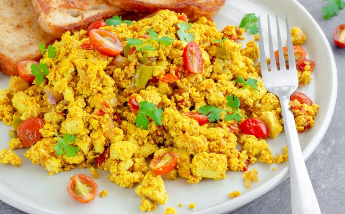 Tofu scramble with tomatoes and vegetables served on a white plate with toast and a fork to illustrate article comparing tofu vs eggs