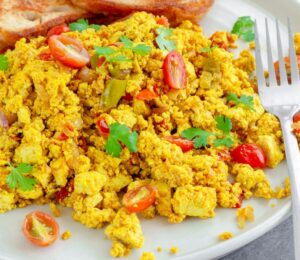Tofu scramble with tomatoes and vegetables served on a white plate with toast and a fork to illustrate article comparing tofu vs eggs