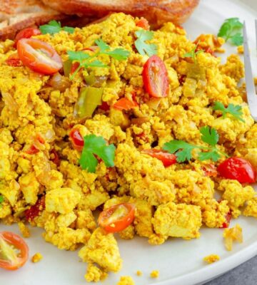 Tofu scramble with tomatoes and vegetables served on a white plate with toast and a fork to illustrate article comparing tofu vs eggs