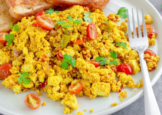 Tofu scramble with tomatoes and vegetables served on a white plate with toast and a fork to illustrate article comparing tofu vs eggs