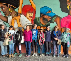Image is a group photo of travelers on one of Ahimsa Travel Club's previous tours. It features a group of people smiling and talking in front of a huge mural. Ahimsa Travel Club could help you take the vegan trip of your dreams