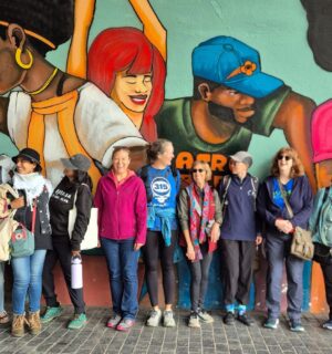 Image is a group photo of travelers on one of Ahimsa Travel Club's previous tours. It features a group of people smiling and talking in front of a huge mural. Ahimsa Travel Club could help you take the vegan trip of your dreams