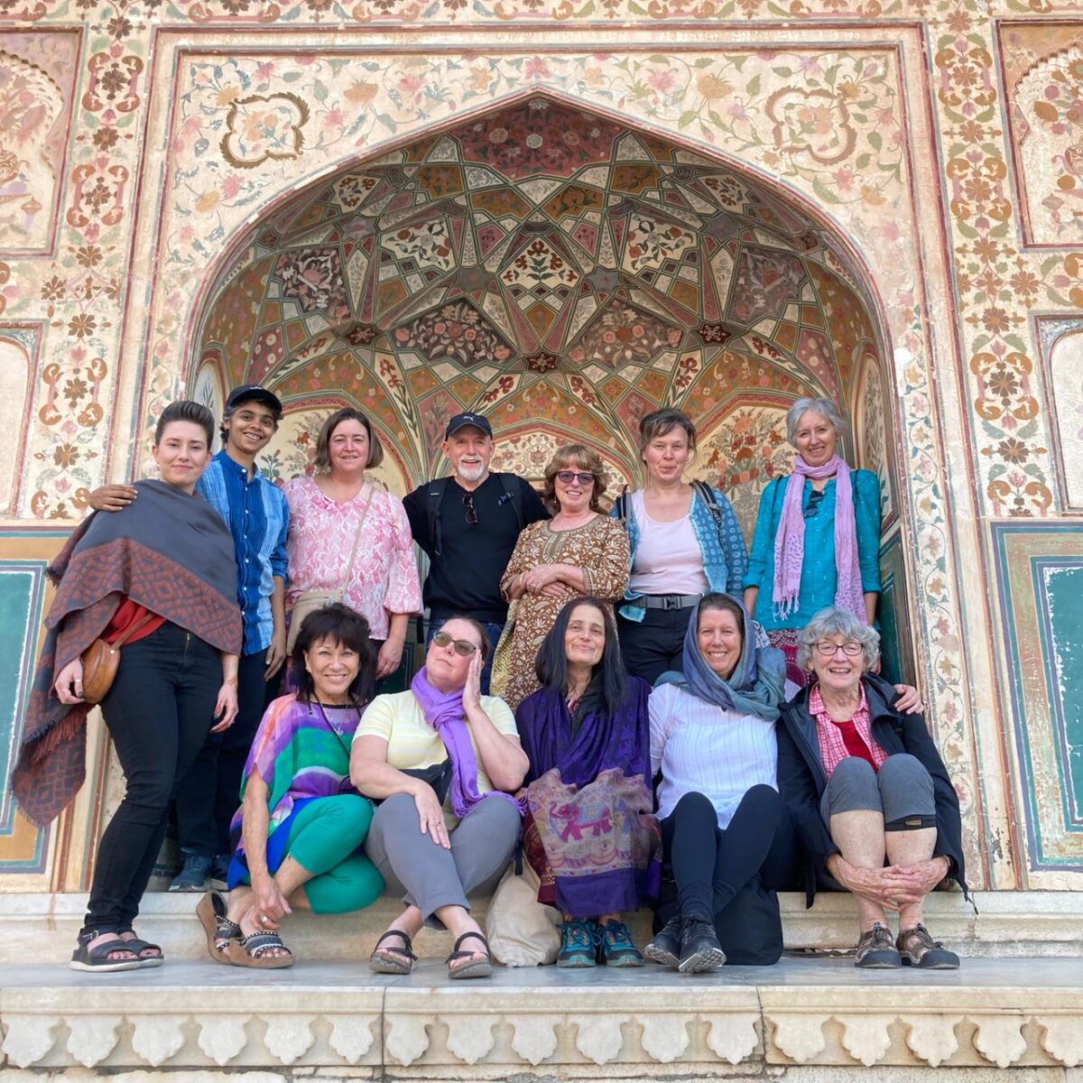Image is a group photo of travelers on one of Ahimsa Travel Club's previous tours. It shows a group of smiling people standing in front of an intricate piece of decorative architecture.