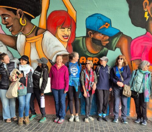 Image is a group photo of travelers on one of Ahimsa Travel Club's previous tours. It features a group of people smiling and talking in front of a huge mural. Ahimsa Travel Club could help you take the vegan trip of your dreams