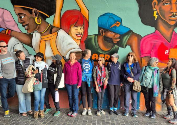 Image is a group photo of travelers on one of Ahimsa Travel Club's previous tours. It features a group of people smiling and talking in front of a huge mural. Ahimsa Travel Club could help you take the vegan trip of your dreams