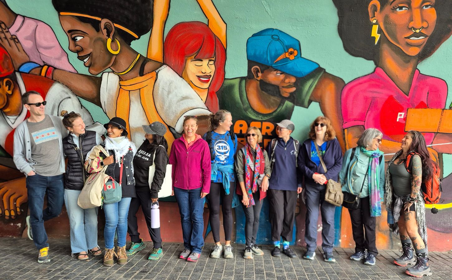 Image is a group photo of travelers on one of Ahimsa Travel Club's previous tours. It features a group of people smiling and talking in front of a huge mural. Ahimsa Travel Club could help you take the vegan trip of your dreams
