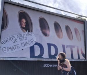 Photo shows a large billboard advertisement for British Airways that has been altered to read, "adverts are flying us into climate chaos," as a young woman walks in front. In a UK first, Bristol City Council has banned high-carbon ads