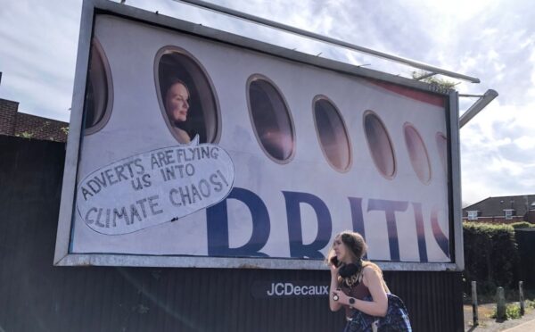 Photo shows a large billboard advertisement for British Airways that has been altered to read, "adverts are flying us into climate chaos," as a young woman walks in front. In a UK first, Bristol City Council has banned high-carbon ads