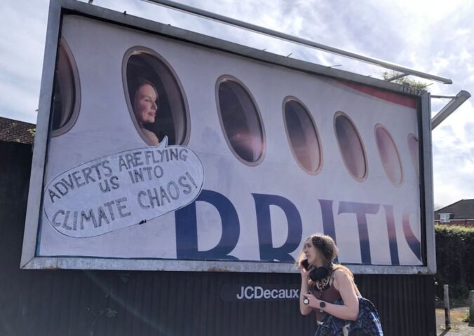 Photo shows a large billboard advertisement for British Airways that has been altered to read, "adverts are flying us into climate chaos," as a young woman walks in front. In a UK first, Bristol City Council has banned high-carbon ads