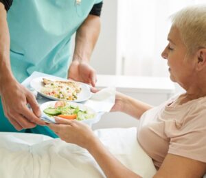Photo shows a woman accepting a tray of plant-based food from staff while laying in a hospital bed. A team of experts has designed a full week of balanced plant-based meals for Dutch hospitals
