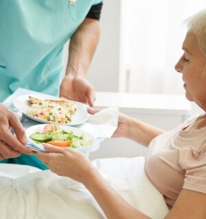 Photo shows a woman accepting a tray of plant-based food from staff while laying in a hospital bed. A team of experts has designed a full week of balanced plant-based meals for Dutch hospitals