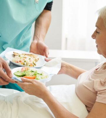 Photo shows a woman accepting a tray of plant-based food from staff while laying in a hospital bed. A team of experts has designed a full week of balanced plant-based meals for Dutch hospitals