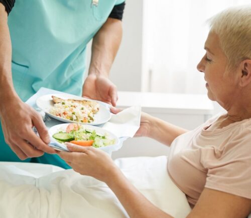 Photo shows a woman accepting a tray of plant-based food from staff while laying in a hospital bed. A team of experts has designed a full week of balanced plant-based meals for Dutch hospitals