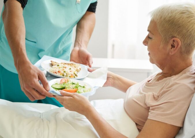Photo shows a woman accepting a tray of plant-based food from staff while laying in a hospital bed. A team of experts has designed a full week of balanced plant-based meals for Dutch hospitals