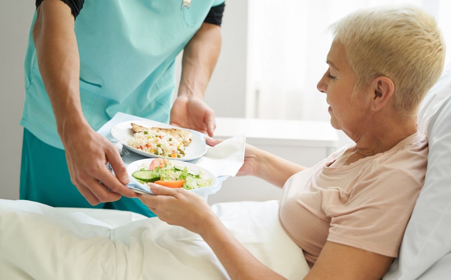 Photo shows a woman accepting a tray of plant-based food from staff while laying in a hospital bed. A team of experts has designed a full week of balanced plant-based meals for Dutch hospitals