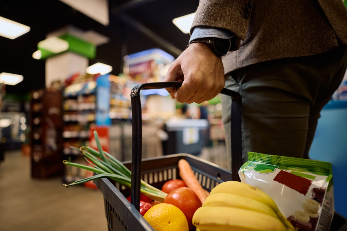Photo shows a man's hand holding a supermarket shopping basket filled with fresh vegetables
