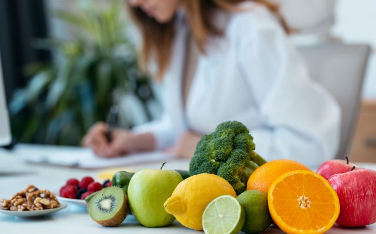 Photo shows a health professional in a white lab coat writing on a clipboard next to a pile of fresh fruit and vegetables. According to new research, most health professionals think that key plant foods are underrepresented in dietary guidelines
