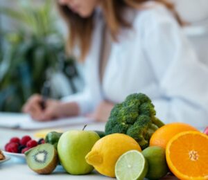 Photo shows a health professional in a white lab coat writing on a clipboard next to a pile of fresh fruit and vegetables. According to new research, most health professionals think that key plant foods are underrepresented in dietary guidelines
