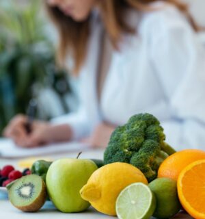 Photo shows a health professional in a white lab coat writing on a clipboard next to a pile of fresh fruit and vegetables. According to new research, most health professionals think that key plant foods are underrepresented in dietary guidelines