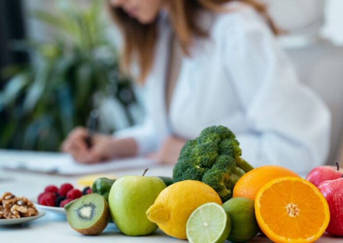 Photo shows a health professional in a white lab coat writing on a clipboard next to a pile of fresh fruit and vegetables. According to new research, most health professionals think that key plant foods are underrepresented in dietary guidelines