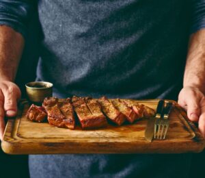 Photo shows a man holding a wooden chopping board with a rare steak, knife, and fork on it. Episode two of Stacey Dooley’s new TV show, Clickbait Clinic, tackles the carnivore diet