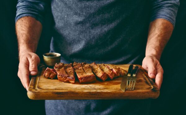 Photo shows a man holding a wooden chopping board with a rare steak, knife, and fork on it. Episode two of Stacey Dooley’s new TV show, Clickbait Clinic, tackles the carnivore diet
