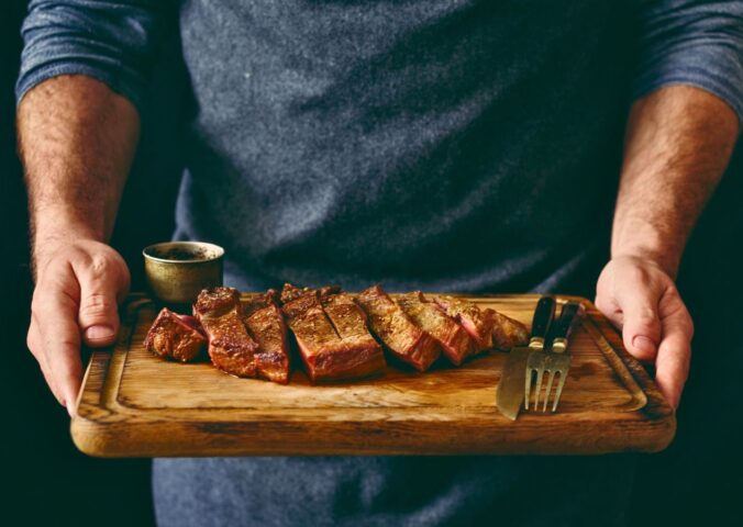 Photo shows a man holding a wooden chopping board with a rare steak, knife, and fork on it. Episode two of Stacey Dooley’s new TV show, Clickbait Clinic, tackles the carnivore diet