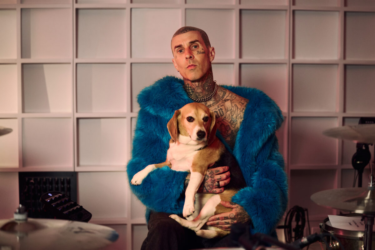 Photo shows Travis Barker holding a rescued dog behind his drum kit while wearing a blue faux fur coat. In a new promotional video for Humane World for Animals, Travis Barker encourages people to “do some good” for animals