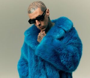 Photo shows Travis Barker wearing sunglasses and a blue faux fur coat. In a new promotional video for Humane World for Animals, Travis Barker encourages people to “do some good” for animals