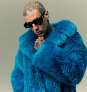 Photo shows Travis Barker wearing sunglasses and a blue faux fur coat. In a new promotional video for Humane World for Animals, Travis Barker encourages people to “do some good” for animals