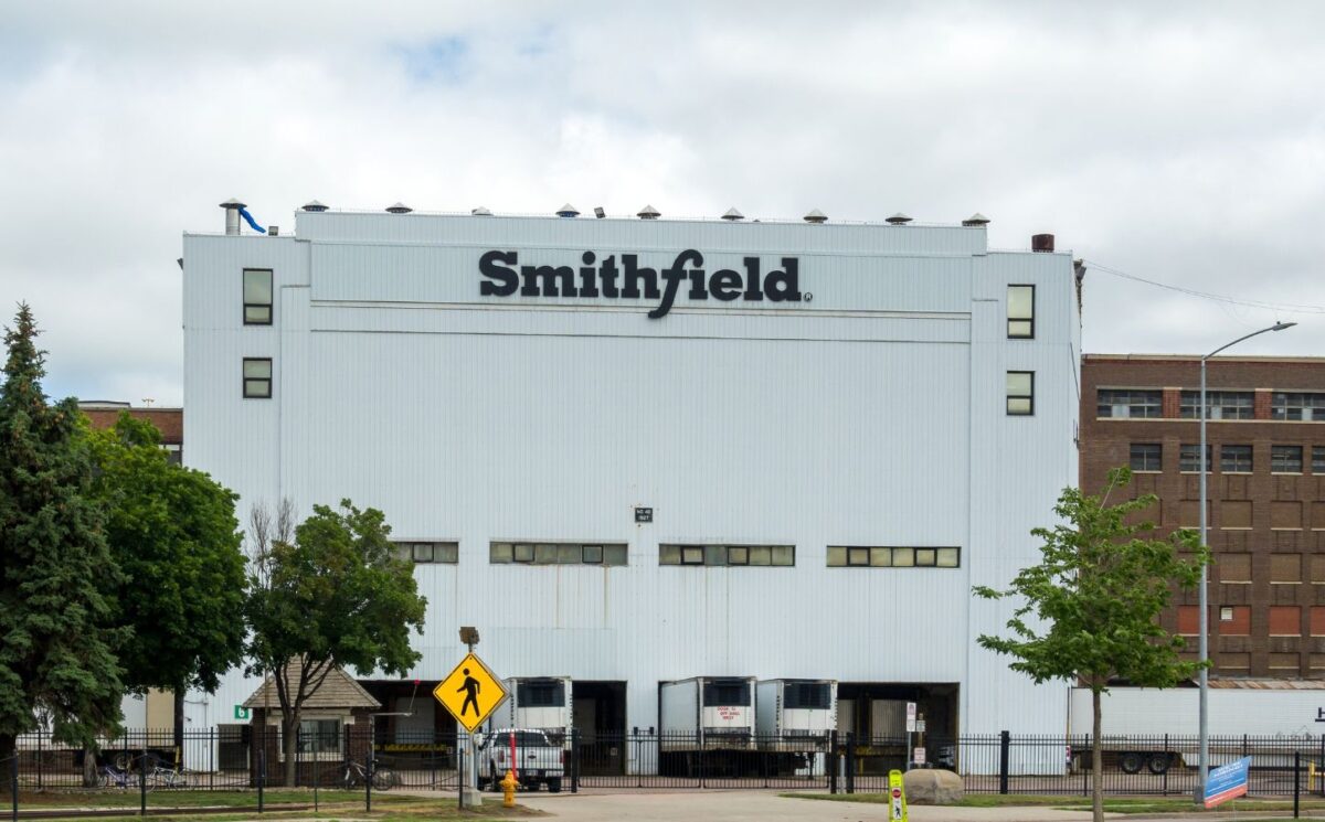 Photo shows the front of a large white Smithfield Foods building, bearing a black "Smithfield" logo. Roughly 100 workers have walked out of a Smithfield meat plant following a colleague’s death