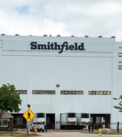 Photo shows the front of a large white Smithfield Foods building, bearing a black "Smithfield" logo. Roughly 100 workers have walked out of a Smithfield meat plant following a colleague’s death