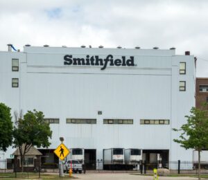 Photo shows the front of a large white Smithfield Foods building, bearing a black "Smithfield" logo. Roughly 100 workers have walked out of a Smithfield meat plant following a colleague’s death