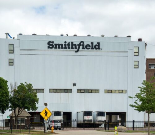 Photo shows the front of a large white Smithfield Foods building, bearing a black "Smithfield" logo. Roughly 100 workers have walked out of a Smithfield meat plant following a colleague’s death