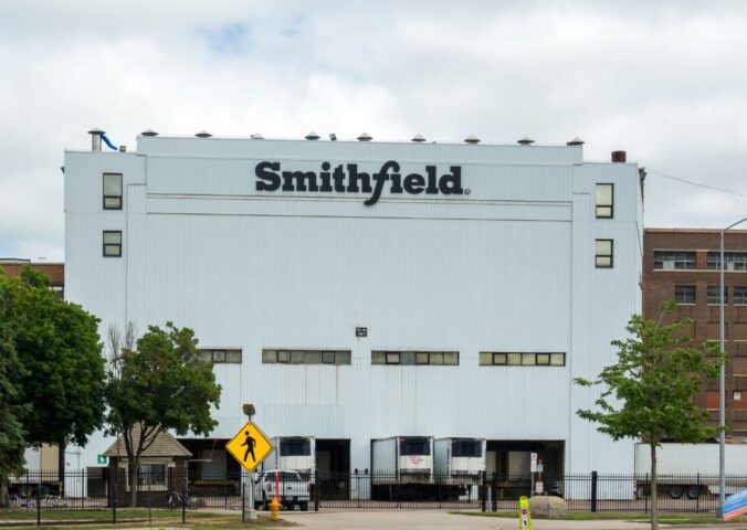 Photo shows the front of a large white Smithfield Foods building, bearing a black "Smithfield" logo. Roughly 100 workers have walked out of a Smithfield meat plant following a colleague’s death